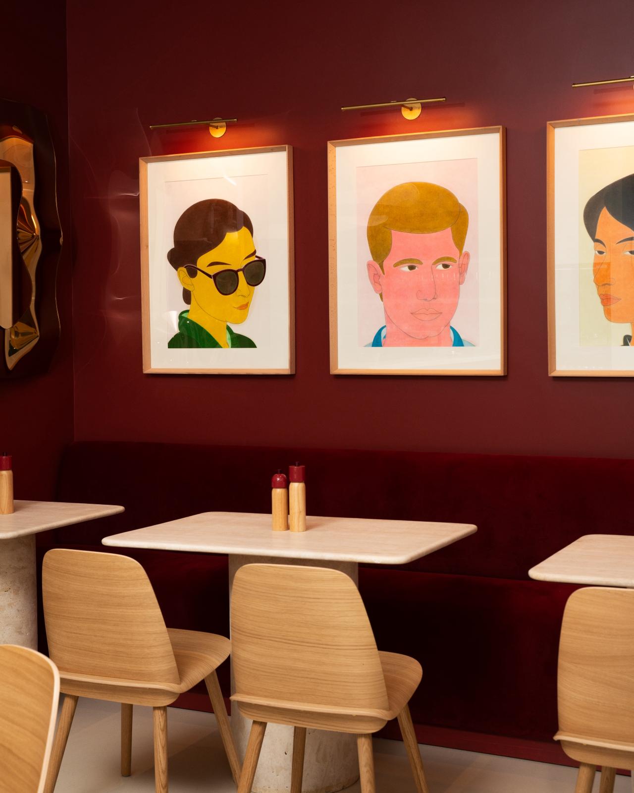 Dajoo interior — oxblood/burgundy wall with three framed colourful Iranian portrait artworks above a burgundy velvet banquette, travertine tables and oak chairs
