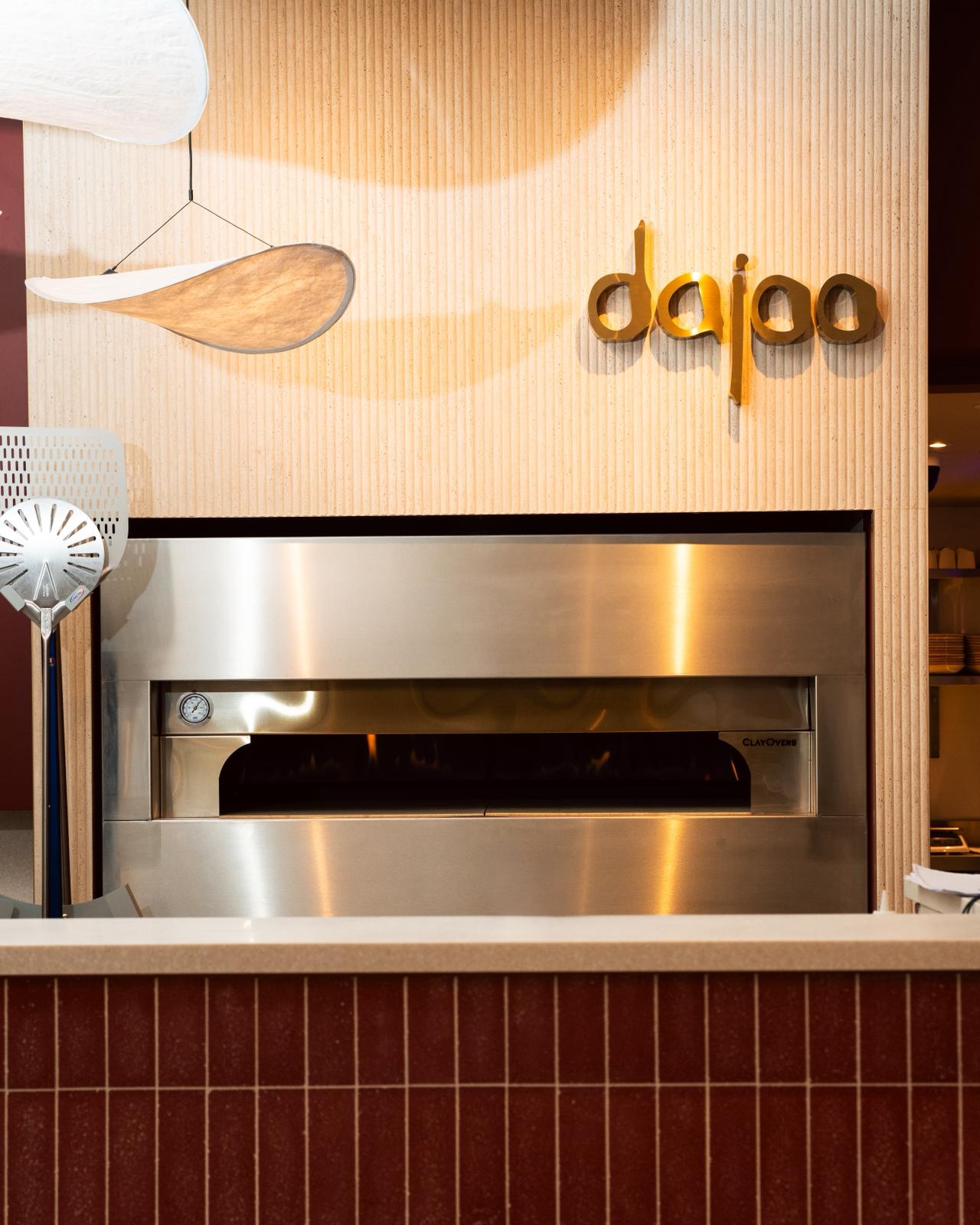 Dajoo clay pizza oven in stainless steel beneath a fluted travertine wall with the gold 3D 'dajoo' sign and a sculptural pendant light