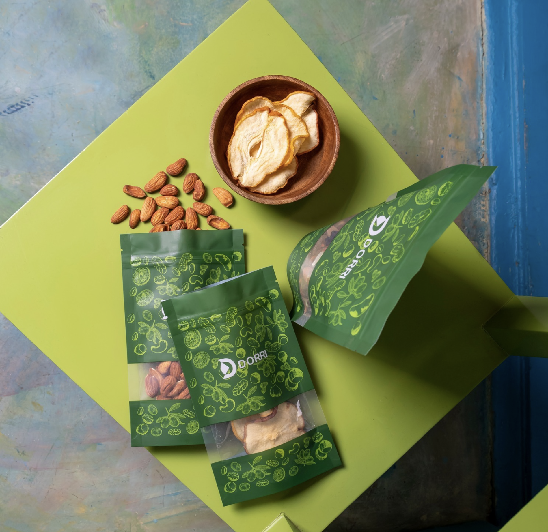 Dorri flat-lay — green botanical-pattern pouches with clear windows, one spilling almonds, beside a wooden bowl of dried pear on a lime board over a painterly blue-grey surface