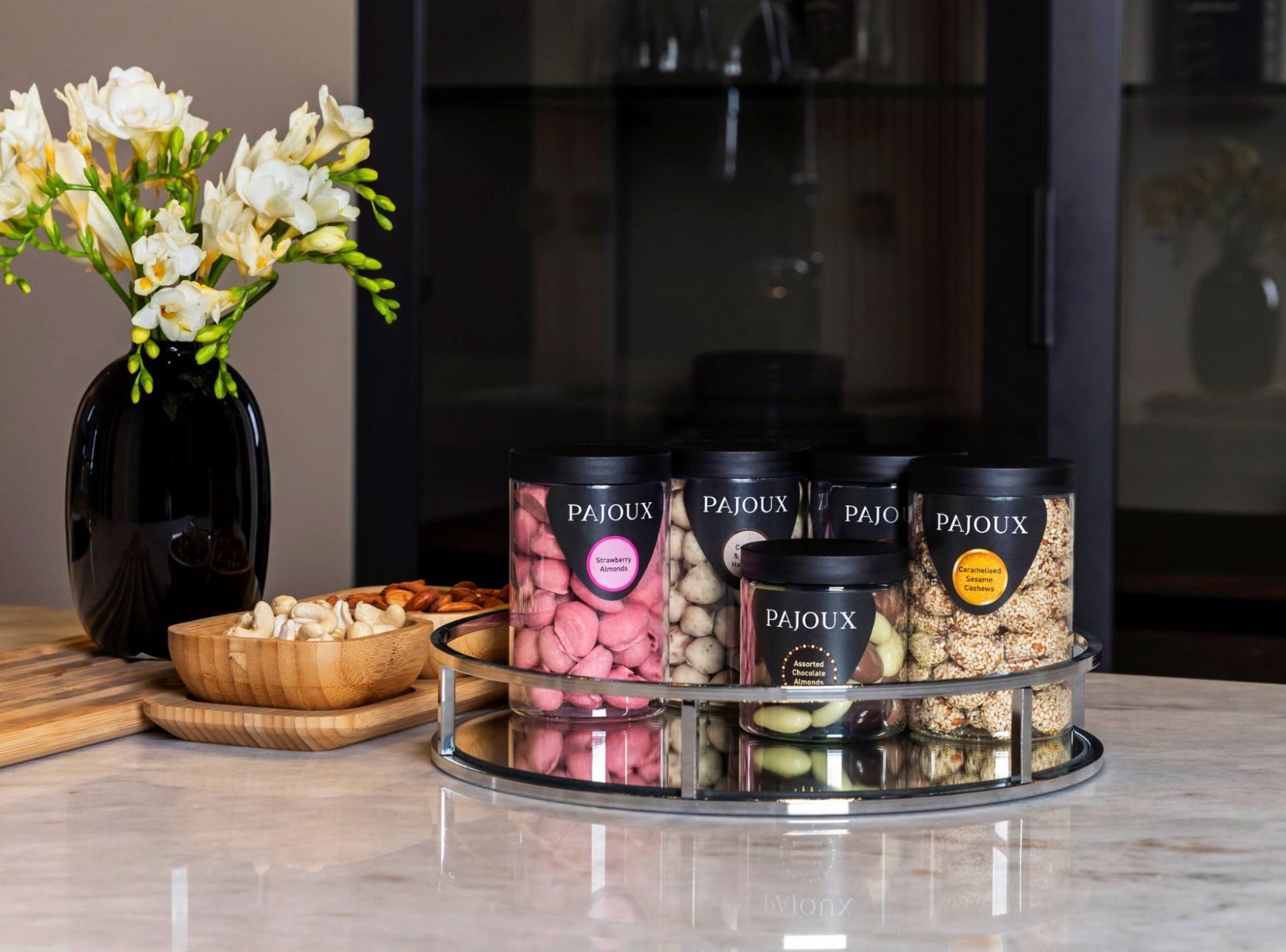 Pajoux kitchen scene — a rotating display of strawberry almonds, sesame cashews and assorted almonds beside white freesias in a black vase on a marble counter