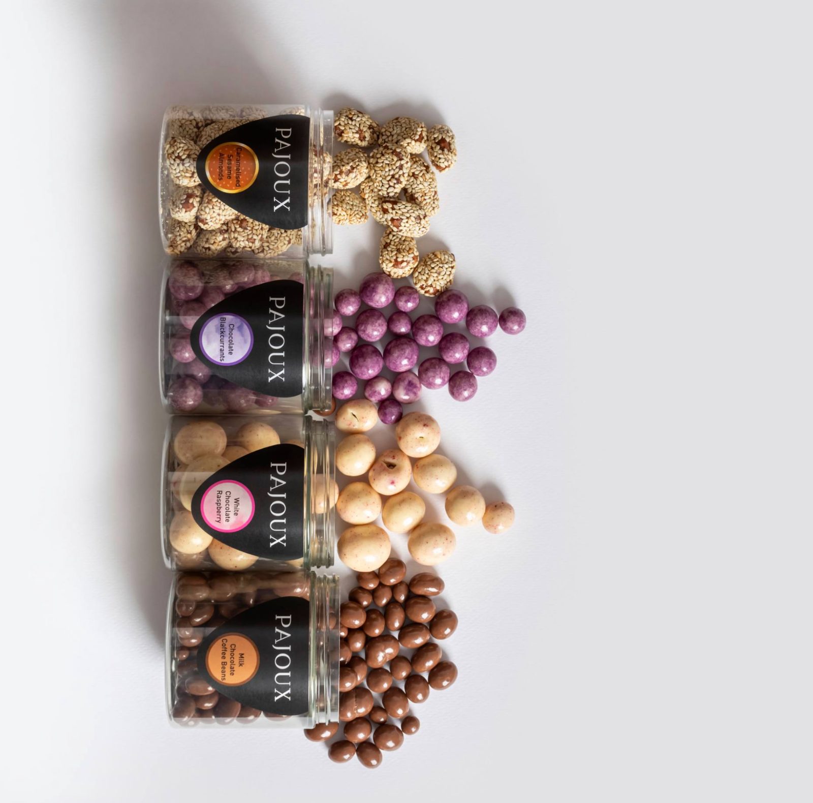 Pajoux four-jar lineup on white — colour-coded badges across sesame almonds, chocolate blackcurrants, white-chocolate raspberry and milk-chocolate coffee beans